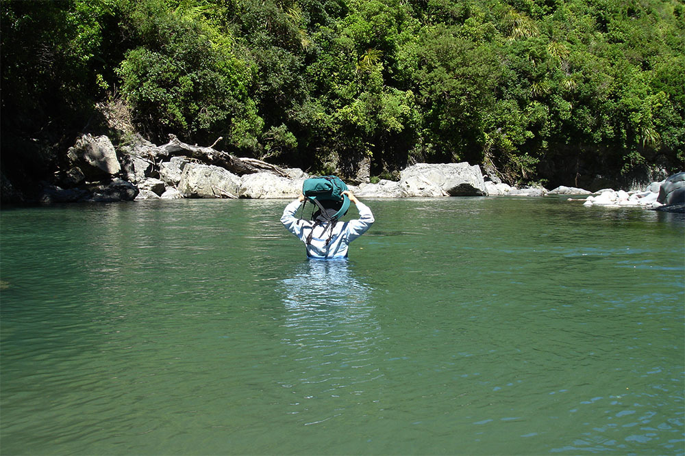 Woman with a rucksack on her head, wading waistdeep in pool of backcountry river, New Zealand